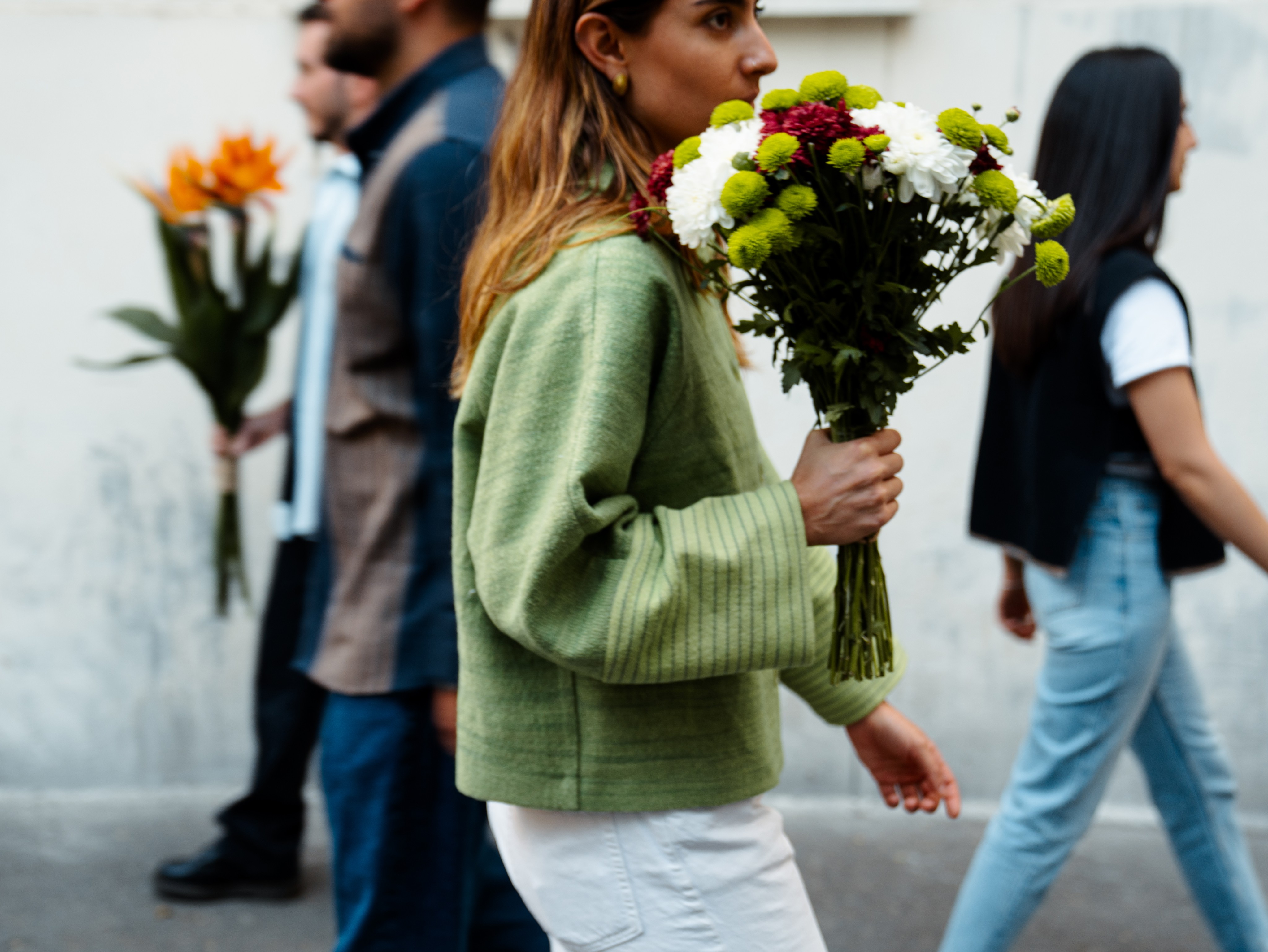 A woman in a sage green jacket carries a bouquet of chrysanthemums past other pedestrians on a pale city street