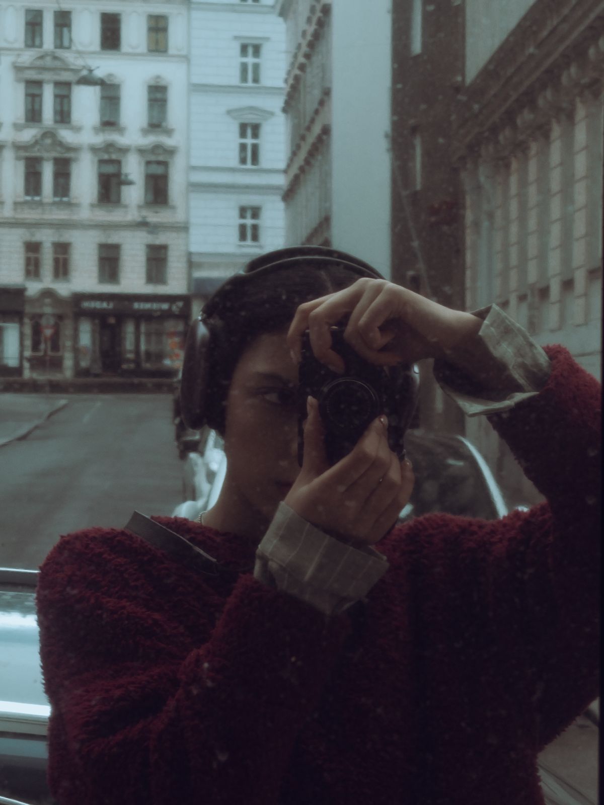 Sara Azadkhani in a window reflection, wearing over-ear headphones and raising a camera to her eye on a European city street
